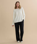A woman with wavy blonde hair stands against a beige background, wearing the Cashmere Chandler Long-Sleeve Tee with a chest pocket, paired with black wide-leg pants and white shoes. Her posture is relaxed and casual.