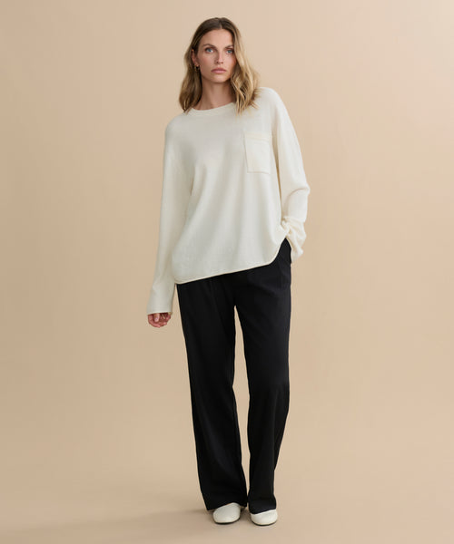 A woman with wavy blonde hair stands against a beige background, wearing the Cashmere Chandler Long-Sleeve Tee with a chest pocket, paired with black wide-leg pants and white shoes. Her posture is relaxed and casual.