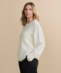 A woman with shoulder-length blonde hair wears the Cashmere Chandler Long-Sleeve Tee featuring a chest pocket, paired with black pants. She stands with one hand in her pocket against a plain beige background.