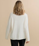 A person with shoulder-length wavy blonde hair is shown from behind, wearing the Cashmere Chandler Long-Sleeve Tee in white and black pants against a beige background.