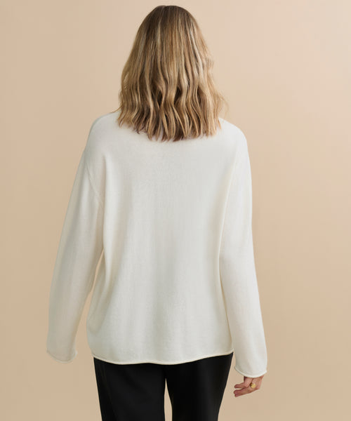 A person with shoulder-length wavy blonde hair is shown from behind, wearing the Cashmere Chandler Long-Sleeve Tee in white and black pants against a beige background.