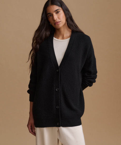 A woman modeling a black ribbed knit cardigan with front buttons over a white top and cream pants, set against a solid beige studio background.