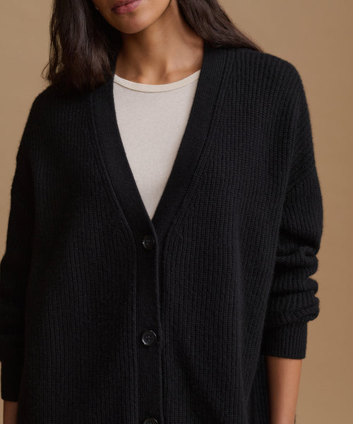 A close-up of a model wearing a black ribbed knit cardigan over a cream-colored crewneck top, set against a solid warm beige background.