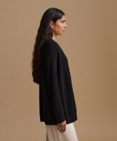 A woman in side profile wearing a black ribbed knit sweater and cream-colored pants, set against a solid warm tan studio background.