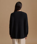 A woman viewed from the back wearing a black ribbed knit sweater and cream-colored pants, set against a solid warm beige studio background.