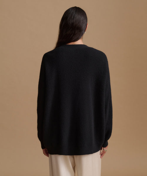 A woman viewed from the back wearing a black ribbed knit sweater and cream-colored pants, set against a solid warm beige studio background.