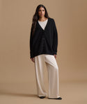 A woman stands against a warm beige studio background wearing a black button-front cardigan over a white top and cream wide-leg pants.