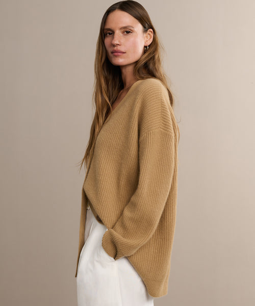 A woman with long brown hair stands against a beige background, wearing the Cashmere Cocoon Cardigan and white pants, looking at the camera with a neutral expression.