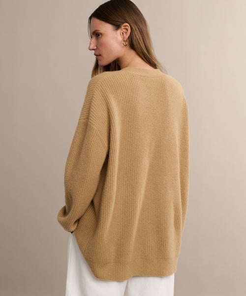 A woman with light brown hair, in the Cashmere Cocoon Cardigan and white pants, stands slightly turned away and looks to her left against a plain beige background.