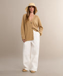 A woman wears a tan wide-brim hat, the Cashmere Cocoon Cardigan, white wide-leg pants, and beige sandals while standing against a neutral background.