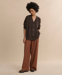 A person stands against a beige background wearing the oversized Cashmere Cocoon Cardigan in dark brown, rust wide-leg trousers, and brown shoes, with one hand in their pocket and looking slightly downward.