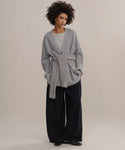 A woman with short curly hair and a calm expression stands against a light beige background, wearing the oversized Cashmere Cocoon Cardigan over a gray top, wide-leg black pants, and black shoes.