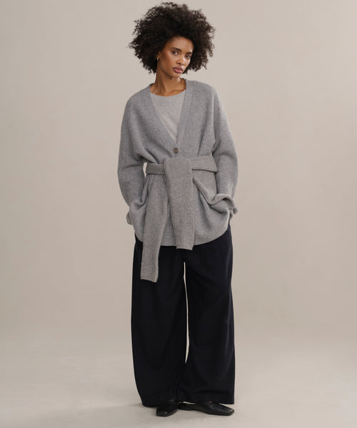 A woman with short curly hair and a calm expression stands against a light beige background, wearing the oversized Cashmere Cocoon Cardigan over a gray top, wide-leg black pants, and black shoes.