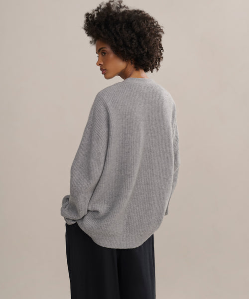 A woman with short curly hair models the oversized gray Cashmere Cocoon Cardigan and black pants, standing with her back turned and glancing over her shoulder against a simple light background.