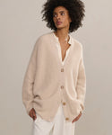 A woman models the Cashmere Cocoon Cardigan in beige over a cream blouse and white pants, standing against a neutral background with her hands in her pockets, gazing to the side.