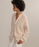 A woman with short curly hair models the Cashmere Cocoon Cardigan over a white blouse and pants, hands in pockets, gazing to the side against a neutral background.