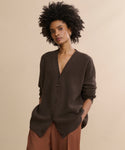 A woman with curly dark hair wears the oversized Cashmere Cocoon Cardigan in dark brown and rust-colored pants, standing against a plain beige background with her hands in her pockets.
