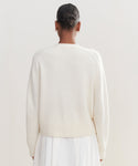 A person with dark hair in a neat bun, wearing small hoop earrings, an ivory Cashmere Colette Cardigan, and a white skirt is shown from the back against a plain light background.
