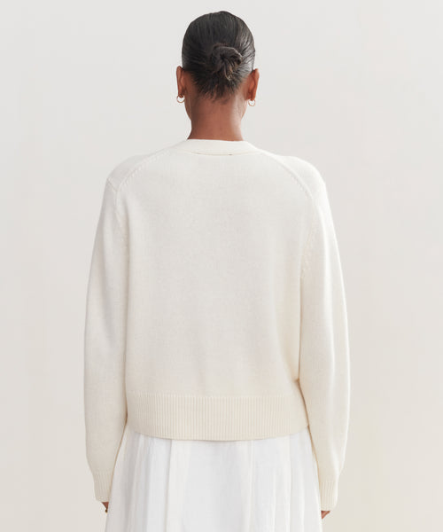 A person with dark hair in a neat bun, wearing small hoop earrings, an ivory Cashmere Colette Cardigan, and a white skirt is shown from the back against a plain light background.