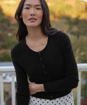 A woman with long dark hair wears the black Cashmere Finley Cardigan, featuring a shrunken fit and button front, paired with a white polka dot skirt while standing outdoors by greenery and a white railing.