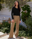 A woman stands on a patio surrounded by greenery, wearing the black Cashmere Finley Cardigan with button front detailing, tan wide-leg pants, and sandals. A white wooden fence and a bench appear in the background.