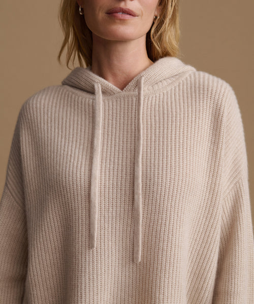 A close-up of a woman wearing a ribbed knit hooded sweater in a light beige color, set against a solid warm tan background.