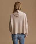 A woman viewed from behind wearing a beige ribbed knit hooded sweater and blue jeans against a solid tan background.