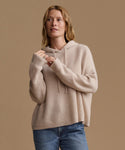 A woman wearing a beige ribbed knit hoodie sweater and blue jeans, posing against a solid warm beige background.