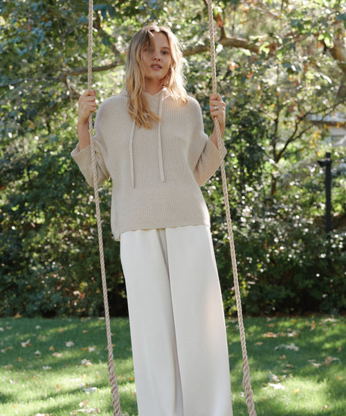 A woman sitting on a rope swing in a garden, wearing a beige ribbed knit sweater and white wide-leg pants.