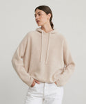 A woman in a light beige Cashmere Fisherman Hoodie with drawstrings and white pants stands against a plain light background, looking to the side with her hands in her pockets.