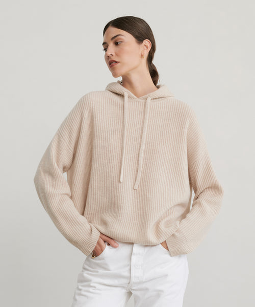 A woman in a light beige Cashmere Fisherman Hoodie with drawstrings and white pants stands against a plain light background, looking to the side with her hands in her pockets.