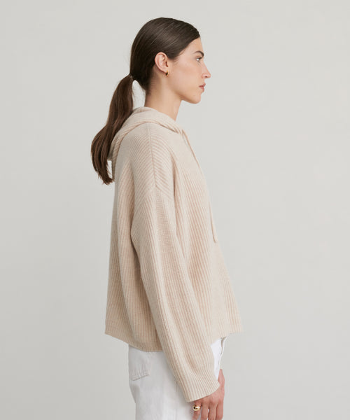 A woman with brown hair in a low ponytail stands in profile against a plain light background, wearing the Cashmere Fisherman Hoodie in oatmeal and white pants.