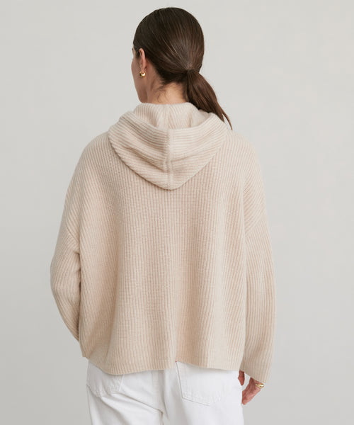 A person with brown hair in a low ponytail, wearing a Cashmere Fisherman Hoodie in oatmeal and white pants, is seen from behind against a plain light background.