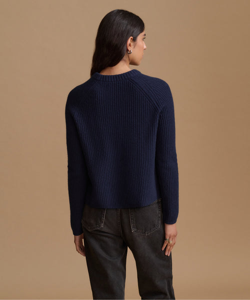 A woman shown from the back wearing a navy blue ribbed knit sweater and dark gray jeans, standing against a solid tan studio background.