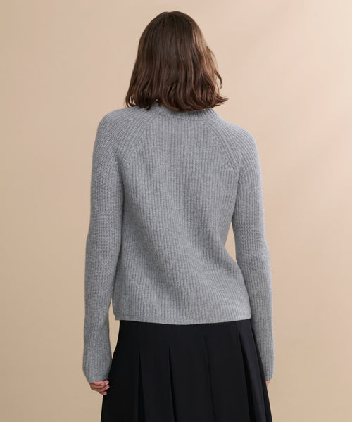 A person with shoulder-length brown hair, seen from behind, wears a heather grey Cashmere Fisherman Sweater and a black skirt against a beige background.
