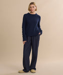 A woman with a blonde bob stands against a beige background wearing the Cashmere Fisherman Sweater in navy, matching loose pants, and beige slippers. She gazes calmly at the camera with one hand in her pocket.