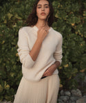 A woman wearing a cream ribbed knit sweater and matching pleated skirt stands outdoors in front of a green hedge.