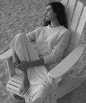 A woman sits barefoot in a white wooden Adirondack chair on sand, wearing a white ribbed knit sweater and matching white skirt. The photo is in black and white.