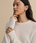 A woman with long dark hair wears a Cashmere Fisherman Sweater in ivory and gold hoop earrings, touching her face thoughtfully to showcase gold rings against a plain beige background.
