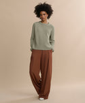 A woman stands against a beige backdrop in a light olive Cashmere Fisherman Sweater and wide brown trousers. With natural curly hair, white shoes, and hands in her pockets, she gazes confidently at the camera.