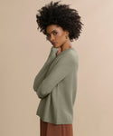 Woman with curly hair stands sideways, arms crossed, wearing the Cashmere Fisherman Sweater in light green and brown pants, looking at the camera against a plain beige background.