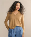 A woman with long curly hair wears the Flynn Cashmere Sweater in camel and light blue pleated pants, standing with one hand in her pocket against a neutral background, smiling subtly.