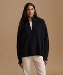A woman modeling a black ribbed knit half-zip sweater with a mock neck, worn over a white top and paired with cream-colored pants, set against a solid tan studio background.