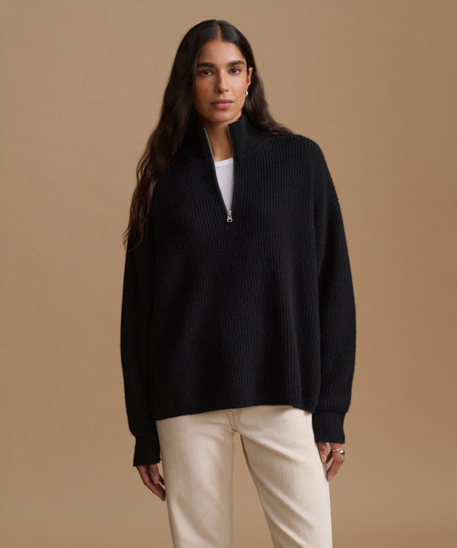 A woman modeling a black ribbed knit half-zip sweater with a mock neck, worn over a white top and paired with cream-colored pants, set against a solid tan studio background.