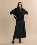 A woman stands against a beige background wearing the Cashmere Half Zip, a long black pleated skirt, and black boots, with her hands on her hips and a neutral expression.