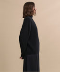 A woman with shoulder-length brown hair stands in profile against a plain beige background, wearing a relaxed Cashmere Half Zip sweater and a long black skirt.