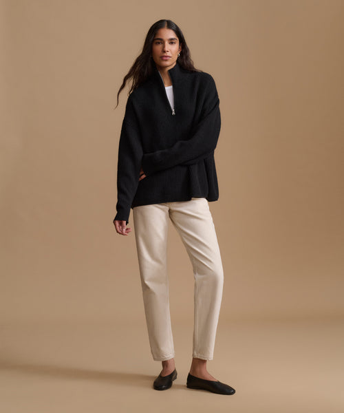 A woman modeling a black long-sleeve zip-front sweater over a white top, paired with cream-colored trousers and black flats, against a solid beige studio background.