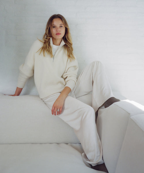 A woman wearing a cream-colored ribbed knit half-zip sweater and matching wide-leg sweatpants, sitting on a white sofa against a white brick wall.