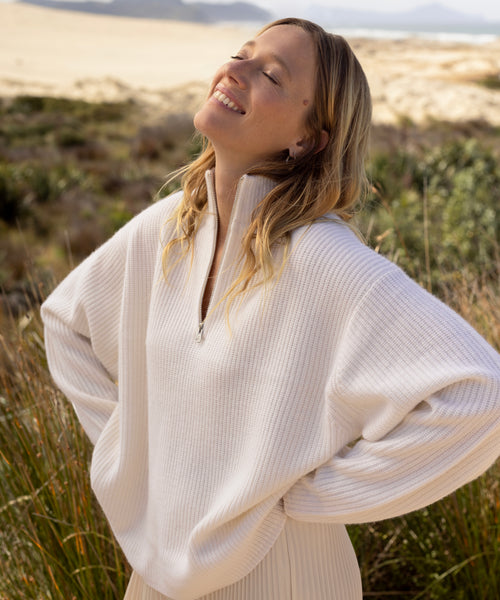 A woman stands outdoors with her eyes closed, head tilted back, and smiling. She wears the Cashmere Half Zip sweater in a grassy, sandy landscape on a sunny day.