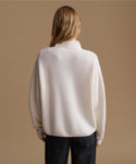 A woman viewed from behind wearing a cream-colored ribbed knit mock-neck sweater and dark trousers, set against a solid warm beige background.
